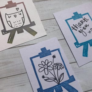 May include: Three small, white cards with illustrations of easels. One card features an owl design, another says "thank you xo", and the third has a floral design. The easel illustrations are in shades of blue, green, and purple.