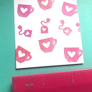 May include: A white card featuring a pattern of pink teacups and tea bags, each with a heart-shaped cutout. A pink ruler with inch measurements is in the foreground, indicating the card's size.