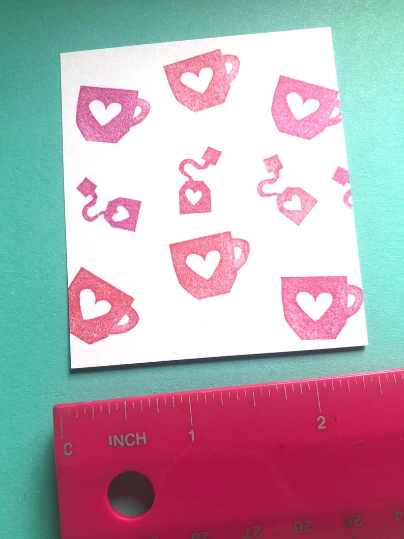 May include: A white card featuring a pattern of pink teacups and tea bags, each with a heart-shaped cutout. A pink ruler with inch measurements is in the foreground, indicating the card's size.