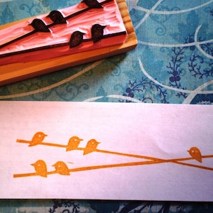 May include: A close-up of a wooden printing block with a design of five birds. The block is used to create a print of birds on a branch, shown in the background.