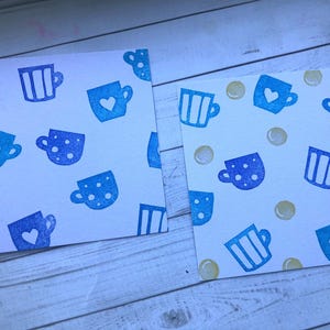 May include: Two white cards with a repeating pattern of blue teacups. The teacups have different designs, including polka dots, stripes, and heart shapes. Some cards also have small yellow circles scattered across the surface.