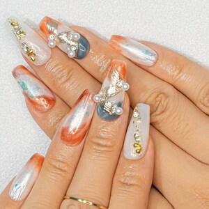 May include: Close-up of hands with long, almond-shaped nails featuring a variety of nail art designs. The nails are painted in shades of white, gray, and orange with intricate details, including gold accents, rhinestones, and pearls.