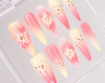 May include: A set of ten false nails in a clear plastic case. The nails are long and pointed, with a gradient of pink and yellow. Some nails have small brown dots and others have a small white flower design.