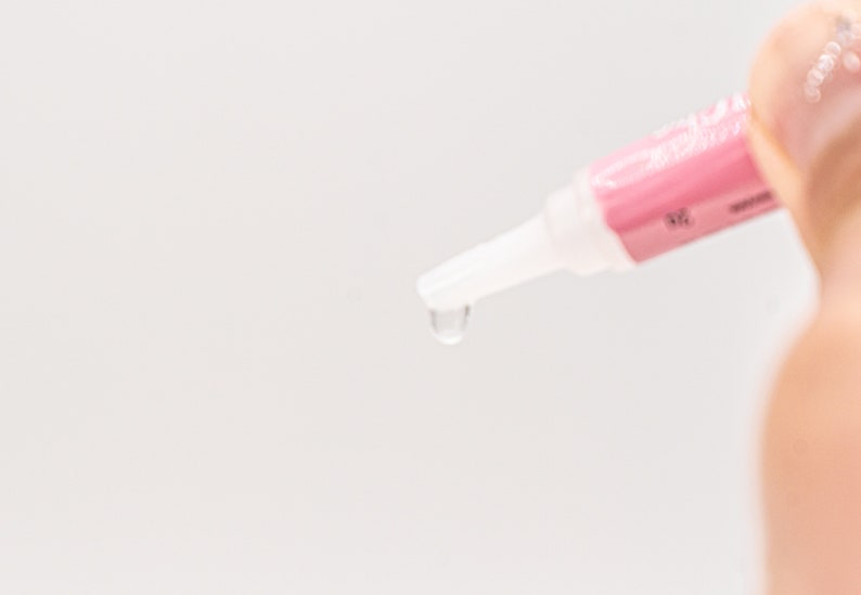 May include: A close-up of a pink and white tube with a clear tip dispensing a drop of liquid.