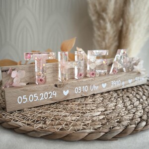 Könnte beinhalten: Ein Holzschild mit dem Namen "THEA" in transparenten Resinbuchstaben, gefüllt mit rosa Blüten und Goldflocken. Das Schild zeigt das Datum 05.05.2024, die Uhrzeit 03:10, das Gewicht 3410g und die Länge 51cm.