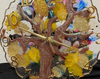 May include: A decorative clock featuring a tree design. The clock face has a clear resin background with a brown tree trunk and gold glitter leaves. Gold numerals mark the hours. The clock is displayed on a black stand.