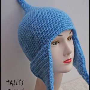 Op de afbeelding: Een lichtblauwe gehaakte muts met oorflappen en een puntige bovenkant. De hoed wordt getoond op een mannequinhoofd. De tekst "Talli's Designs" is zichtbaar in de linker benedenhoek.