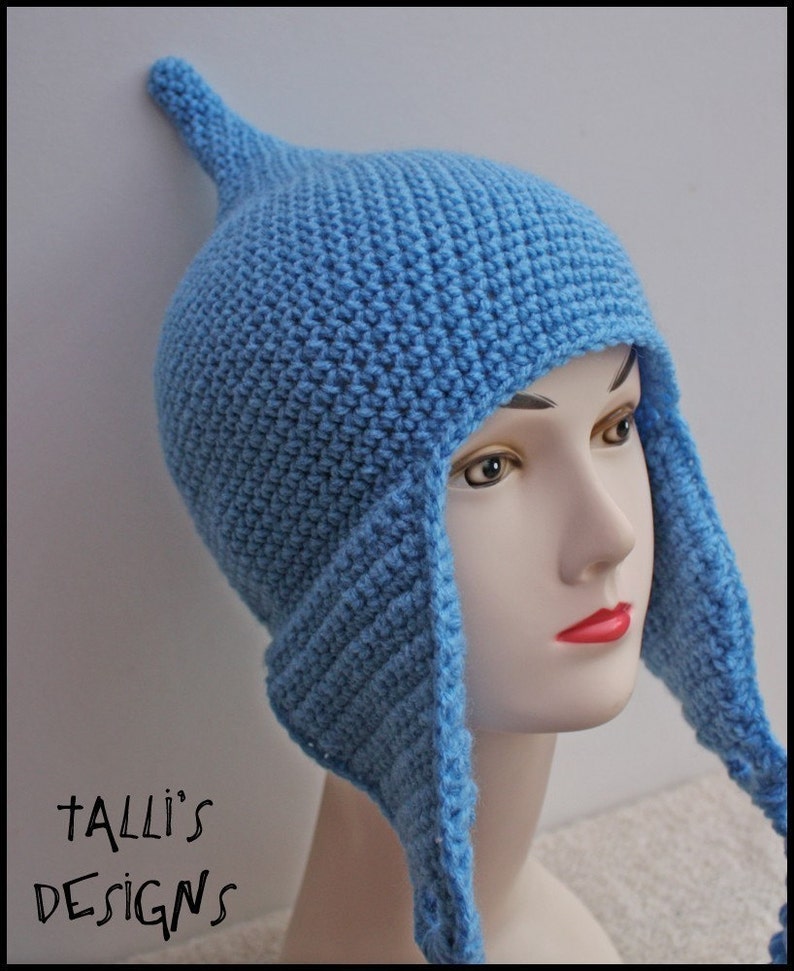 Op de afbeelding: Een lichtblauwe gehaakte muts met oorflappen en een puntige bovenkant. De hoed wordt getoond op een mannequinhoofd. De tekst "Talli's Designs" is zichtbaar in de linker benedenhoek.