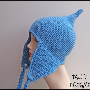 Peut inclure: Un bonnet bleu clair en crochet avec un sommet pointu et des rabats pour les oreilles. Le bonnet a des liens tressés et est présenté sur une tête de mannequin. Le texte "Talli's Designs" est visible dans le coin inférieur droit.