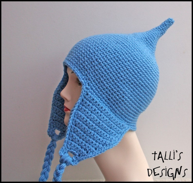 Op de afbeelding: Een lichtblauwe gehaakte hoed met een puntige top en oorflappen. De hoed heeft gevlochten banden en wordt getoond op een mannequinhoofd. De tekst "Talli's Designs" is zichtbaar in de rechter benedenhoek.
