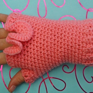 May include: A coral-colored, crocheted fingerless glove with a ruffled accent. The glove is made of a textured yarn and covers the hand up to the knuckles and wrist. The background is a light blue with pink swirls.