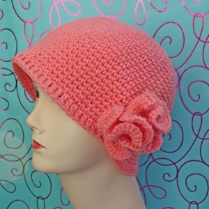 May include: A coral-colored, crocheted hat with a small brim and a large, decorative flower on the side. The hat is displayed on a mannequin head. The hat is handmade and appears to be made of soft yarn.