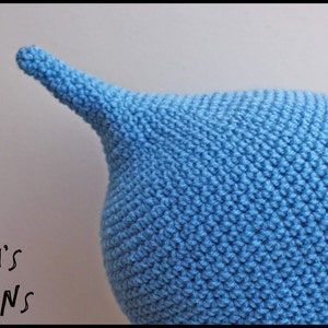 Op de afbeelding: Een close-up van een lichtblauwe gehaakte muts met een puntige top. De muts is handgemaakt met een gestructureerd steekpatroon. De tekst "Talli's Designs" staat in de linkerbenedenhoek. De muts is waarschijnlijk voor een baby of kind.