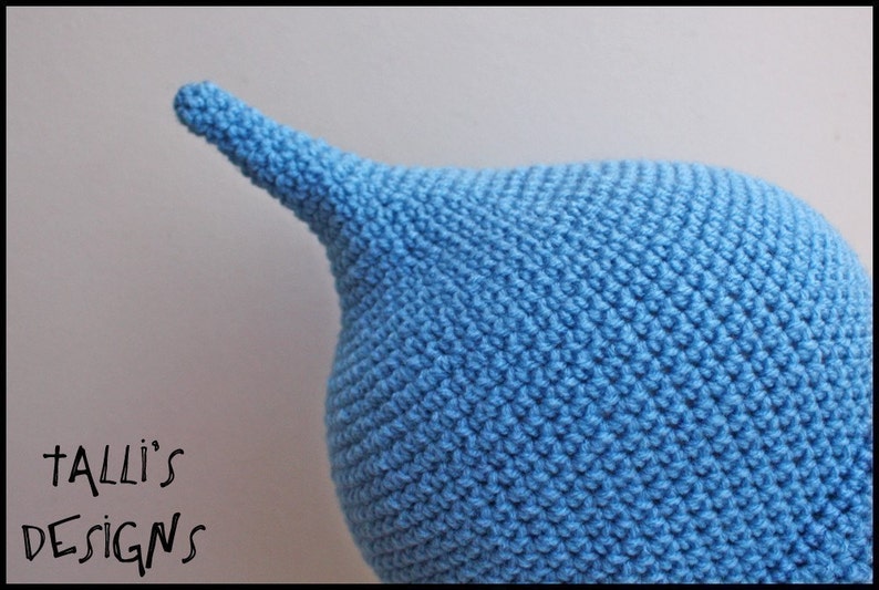 Op de afbeelding: Een close-up van een lichtblauwe gehaakte muts met een puntige top. De muts is handgemaakt met een gestructureerd steekpatroon. De tekst "Talli's Designs" staat in de linkerbenedenhoek. De muts is waarschijnlijk voor een baby of kind.