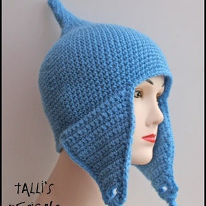 Op de afbeelding: Een lichtblauwe gehaakte muts met oorflappen en een puntige bovenkant. De hoed wordt getoond op een mannequinhoofd. De tekst "Talli's Designs" is zichtbaar in de linker benedenhoek.