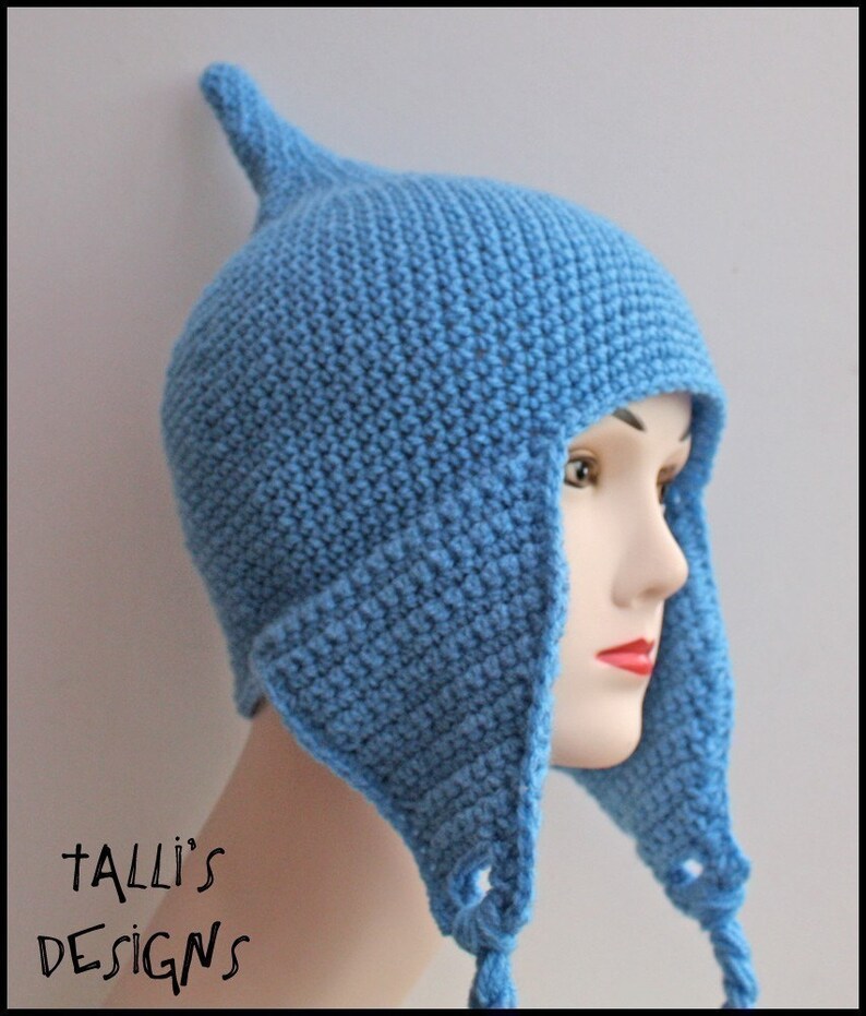 Op de afbeelding: Een lichtblauwe gehaakte muts met oorflappen en een puntige bovenkant. De hoed wordt getoond op een mannequinhoofd. De tekst "Talli's Designs" is zichtbaar in de linker benedenhoek.