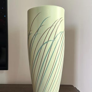 Può includere: Un vaso alto in ceramica verde chiaro con un disegno di erba stilizzata marrone e blu.