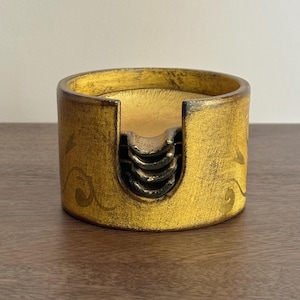 May include: A set of four round wooden coasters with a gold finish and a black floral design. The coasters are stacked inside a matching wooden holder.