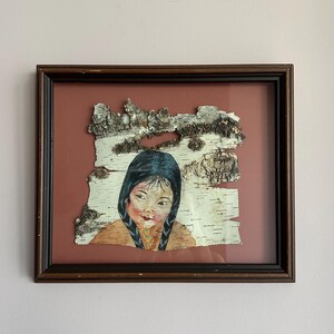May include: Framed artwork featuring a portrait of a young girl with dark braids, painted on a piece of birch bark. The artwork is set against a reddish-brown background within a dark brown wooden frame. The girl's face is painted with soft colors.