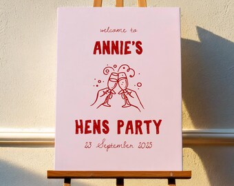 Könnte beinhalten: Ein rosafarbenes Willkommenszeichen mit dem Text "welcome to ANNIE'S HENS PARTY 23 September 2025" in roten Buchstaben. Das Schild zeigt eine Grafik von zwei Champagnergläsern, die anstoßen.