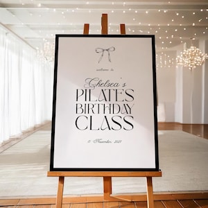 May include: A framed sign on a wooden easel. The sign reads "Chelsea's Pilates Birthday Class" with the date "11 November 2021" below. A bow graphic is at the top. The background is a bright room with white curtains and a chandelier.