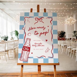 May include: A wooden easel displays a baby shower sign with a blue and white checkered border. The sign reads "She's ready to pop!" with a red Coca-Cola bottle illustration and the text "Welcome to Hannah's Baby Shower 16 July 2027."