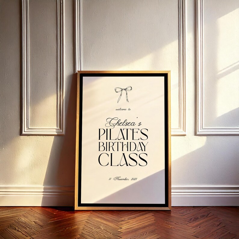May include: A framed poster with a gold border, featuring the text "welcome to Chelsea's PILATES BIRTHDAY CLASS" and the date "11 November 2021". A bow graphic is at the top. The poster leans against a white wall.