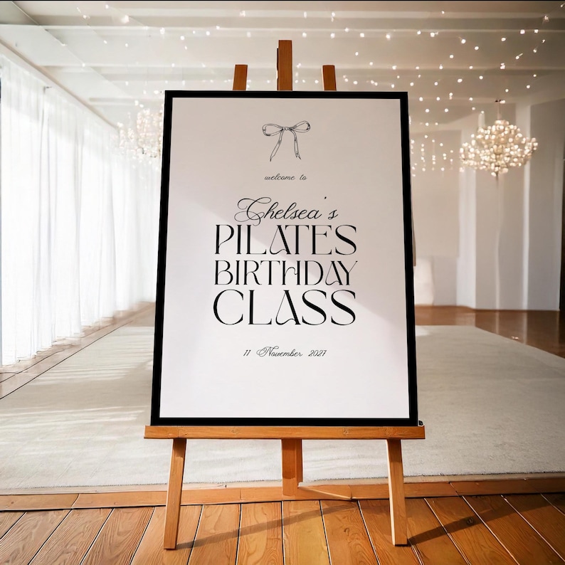 May include: A framed sign on a wooden easel. The sign reads "Chelsea's Pilates Birthday Class" with the date "11 November 2021" below. A bow graphic is at the top. The background is a bright room with white curtains and a chandelier.