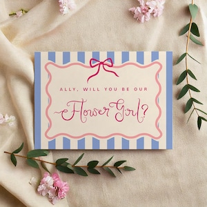 以下が含まれることがあります： ピンク色の筆記体で「ALLY, WILL YOU BE OUR Flower Girl?」と書かれた、花をテーマにした招待状。カードは、ピンクのリボンとボーダーが付いた、水色と白のストライプの背景です。装飾的な花と緑がカードを囲んでいます。