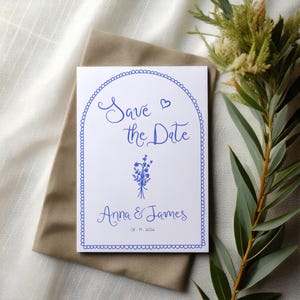 May include: A white save the date card with a blue border and a blue floral illustration. The text reads "Save the Date" with a heart symbol and "Anna & James" with the date "08.11.2026".