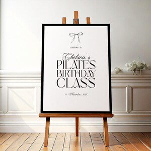 May include: A framed sign on a wooden easel with the text "Chelsea's Pilates Birthday Class" in elegant black script. The sign includes a bow illustration and the date "11 November 2021". The background features a white wall and wooden floor.