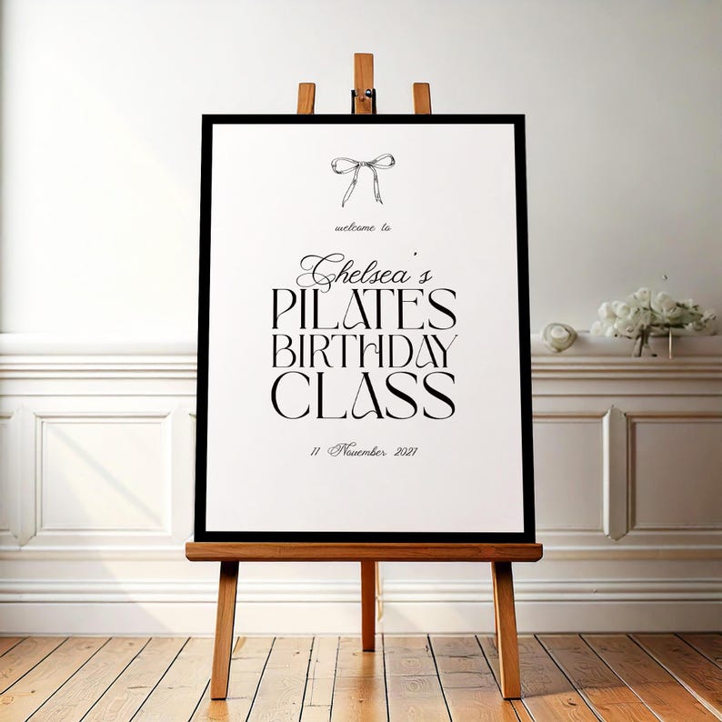 May include: A framed sign on a wooden easel with the text "Chelsea's Pilates Birthday Class" in elegant black script. The sign includes a bow illustration and the date "11 November 2021". The background features a white wall and wooden floor.
