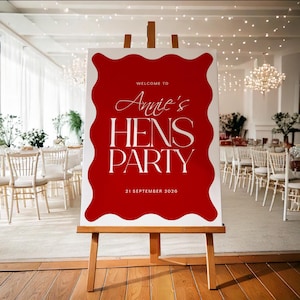 Puede incluir: Un caballete de madera exhibe un letrero con fondo rojo y borde blanco ondulado. El letrero dice "Welcome to Annie's HENS PARTY" en elegante escritura blanca y letras mayúsculas, con la fecha "21 de septiembre de 2020" en la parte inferior.