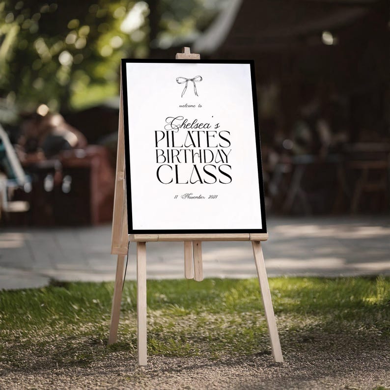 May include: A white sign with black text on a wooden easel. The sign reads "Chelsea's Pilates Birthday Class" with a bow illustration at the top. The date "11 November 2021" is at the bottom. The easel is set on grass.