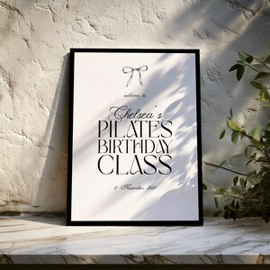 May include: A framed print with the text "welcome to Chelsea's PILATES BIRTHDAY CLASS" in black. A bow illustration is at the top, and the date "11 November 2021" is at the bottom. The print is displayed on a marble surface.