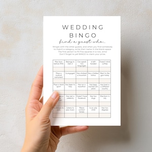 Op de afbeelding: Een witte trouwbingokaart met zwarte tekst. De kaart, die door een hand wordt vastgehouden, heeft een raster met prompts om gasten te vinden die bij de beschrijvingen passen. De bovenkant leest "WEDDING BINGO".