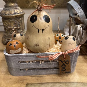 May include: A gray wooden crate filled with wood shavings containing a large central gourd painted as a ghost with fangs, two small painted pumpkin gourds, and one small ghost gourd. A wooden tag with the word Boo! is tied to the crate with ribbon.