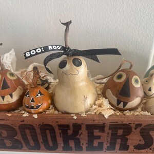 May include: A rectangular wooden crate labeled BOOKER'S containing several painted gourd decorations. The gourds feature hand-painted faces, including jack-o'-lanterns and a ghost-like figure with a black ribbon tied at the top reading BOO!.