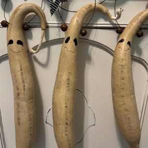 May include: Three gourd-shaped Halloween decorations with smiling faces and curved stems. The gourds are painted with black eyes and mouths. They hang from a wooden rod decorated with black and white striped ribbon and black accents.