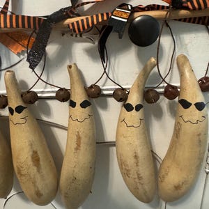 May include: A decorative garland featuring four light tan, elongated gourd ornaments hanging from a wooden rod. Each gourd is hand-painted with black eyes and a wavy mouth. The arrangement is accented with orange and black striped ribbons.