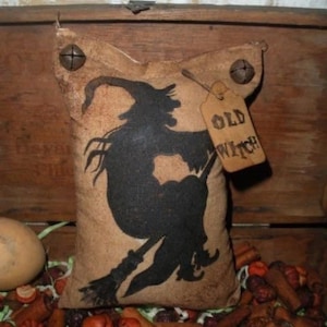 May include: A tan fabric pillow with a black silhouette of a witch on a broomstick. The pillow has a tag that reads "OLD WITCH". Two metal fasteners are attached to the top corners. The pillow is surrounded by fall-themed decorations.