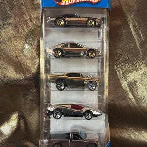Op de afbeelding: Een Hot Wheels Shiners 5-pack speelgoedauto's in een blauwe en transparante plastic verpakking. De auto's zijn in verschillende tinten metallic goud, zilver en bruin. De verpakking heeft het Hot Wheels-logo en de tekst "Shiners".