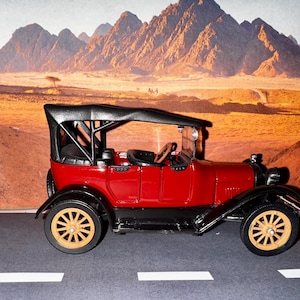 Könnte beinhalten: Ein rotes Vintage-Automodell mit schwarzem Stoffverdeck und Holzrädern. Das Auto ist vor einem Wüstenhintergrund mit Bergen in der Ferne platziert.