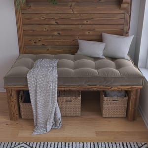 Op de afbeelding: Een houten bank met een getuft taupekleurig kussen, twee lichtgrijze sierkussens en een gestructureerde grijze plaid die over de zijkant hangt. Drie gevlochten rieten opbergmanden staan onder de bank op de houten vloer.