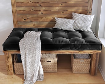 May include: A wooden bench featuring a thick black tufted cushion, two white decorative pillows with textured geometric patterns, and a light gray knit throw blanket draped over the side. Three woven wicker storage baskets sit underneath.