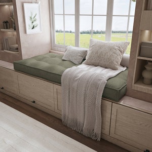 May include: A green window seat cushion with a white throw blanket and a white pillow. The window seat is built into a set of wooden cabinets.