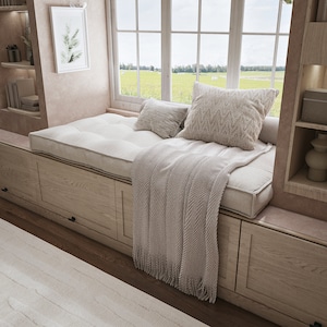May include: A cozy window seat with a tufted cushion, two decorative pillows, and a fringed throw blanket. The seat is built into a wooden cabinet with storage drawers. A framed botanical print hangs on the wall, and a large window overlooks a green field.
