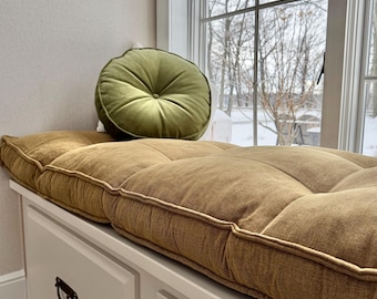 May include: A long, rectangular, tufted mustard-yellow bench cushion with a textured fabric finish, paired with a round, olive-green velvet throw pillow featuring a central button tuft, both resting on a white wooden storage bench.