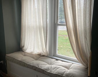 May include: A white window seat bench featuring a tufted, beige rectangular cushion. The bench is positioned beneath a window framed by two light beige, semi-sheer curtains hanging from a gold-toned metal curtain rod against a dark wall.