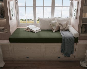 May include: A cozy window seat with a dark green cushion, white pillows, and a rolled bolster. An open book rests on the cushion, and a gray and white striped throw blanket is draped over the side. The window offers a view of a blue sky and green landscape.